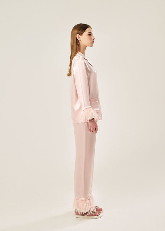 Side view of a blush pink viscose mix rayon pajama set featuring a classic collar, floral buttons, and two front pockets. The detachable ostrich feather trim on the cuffs and pant hems adds a luxurious touch to the relaxed silhouette.