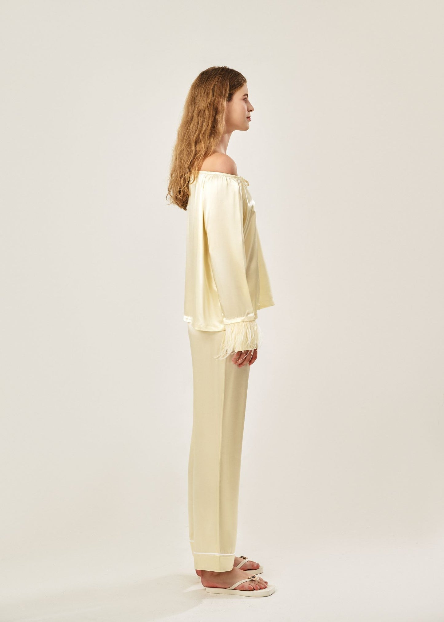 Side view of a beige off-shoulder pajama set in soft viscose with a silky feel, featuring a delicate bow detail on the neckline and waistband. Detachable ostrich feather trim on the sleeves adds a chic, elegant finish to the relaxed silhouette.