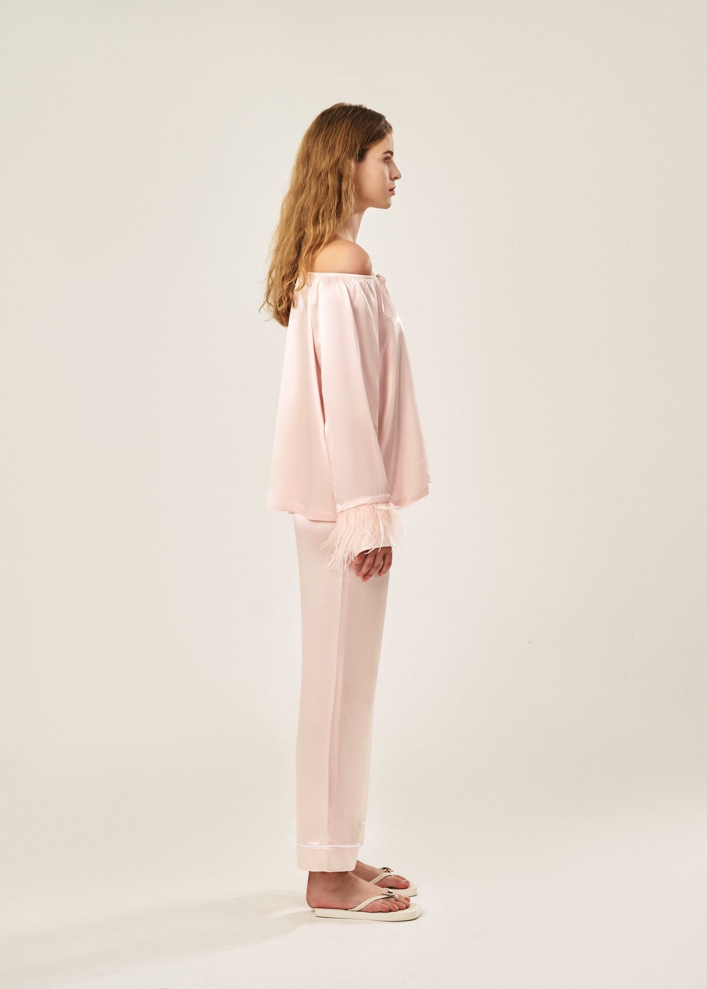 Side view of a blush pink off-shoulder pajama set in soft viscose with a silky feel, featuring a delicate bow detail on the neckline and waistband. Detachable ostrich feather trim on the sleeves adds a chic, elegant finish to the relaxed silhouette.