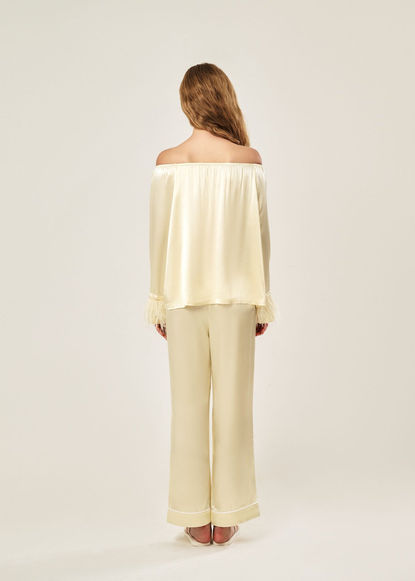 Back view of a beige off-shoulder pajama set in soft viscose with a silky feel, featuring a delicate bow detail on the neckline and waistband. Detachable ostrich feather trim on the sleeves adds a chic, elegant finish to the relaxed silhouette.