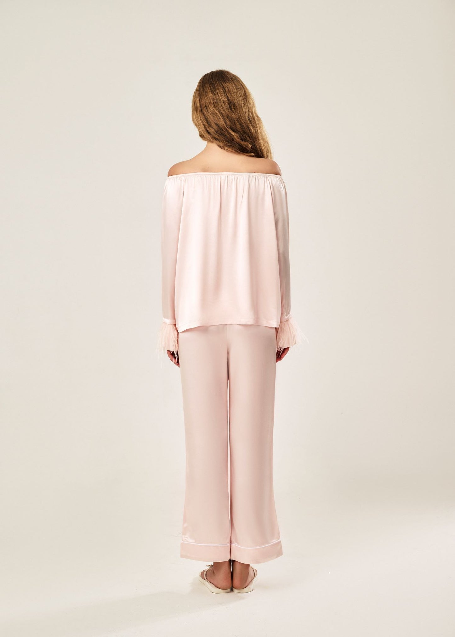 Back view of a blush pink off-shoulder pajama set in soft viscose with a silky feel, featuring a delicate bow detail on the neckline and waistband. Detachable ostrich feather trim on the sleeves adds a chic, elegant finish to the relaxed silhouette.
