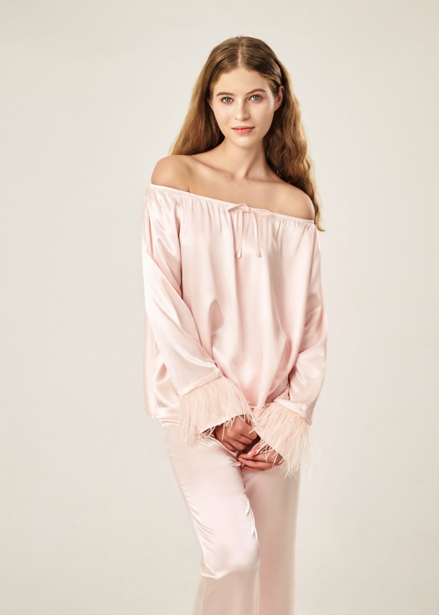 Detail view of a blush pink off-shoulder pajama set in soft viscose with a silky feel, featuring a delicate bow detail on the neckline and waistband. Detachable ostrich feather trim on the sleeves adds a chic, elegant finish to the relaxed silhouette.