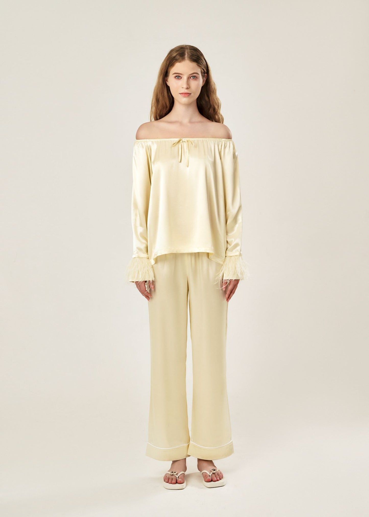 Front view of a beige off-shoulder pajama set in soft viscose with a silky feel, featuring a delicate bow detail on the neckline and waistband. Detachable ostrich feather trim on the sleeves adds a chic, elegant finish to the relaxed silhouette.