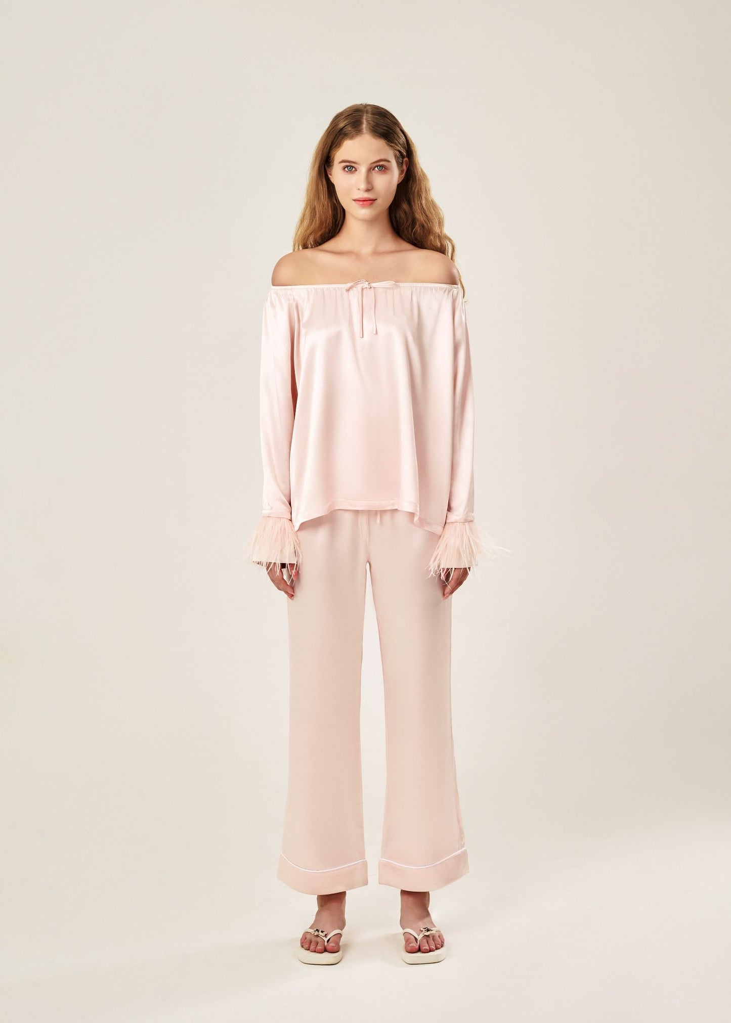 Front view of a blush pink off-shoulder pajama set in soft viscose with a silky feel, featuring a delicate bow detail on the neckline and waistband. Detachable ostrich feather trim on the sleeves adds a chic, elegant finish to the relaxed silhouette.