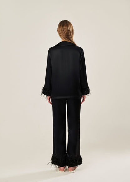 Back view of black viscose mix rayon pajama set featuring a classic collar, floral buttons, and two front pockets. The detachable ostrich feather trim on the cuffs and pant hems adds a luxurious touch to the relaxed silhouette.