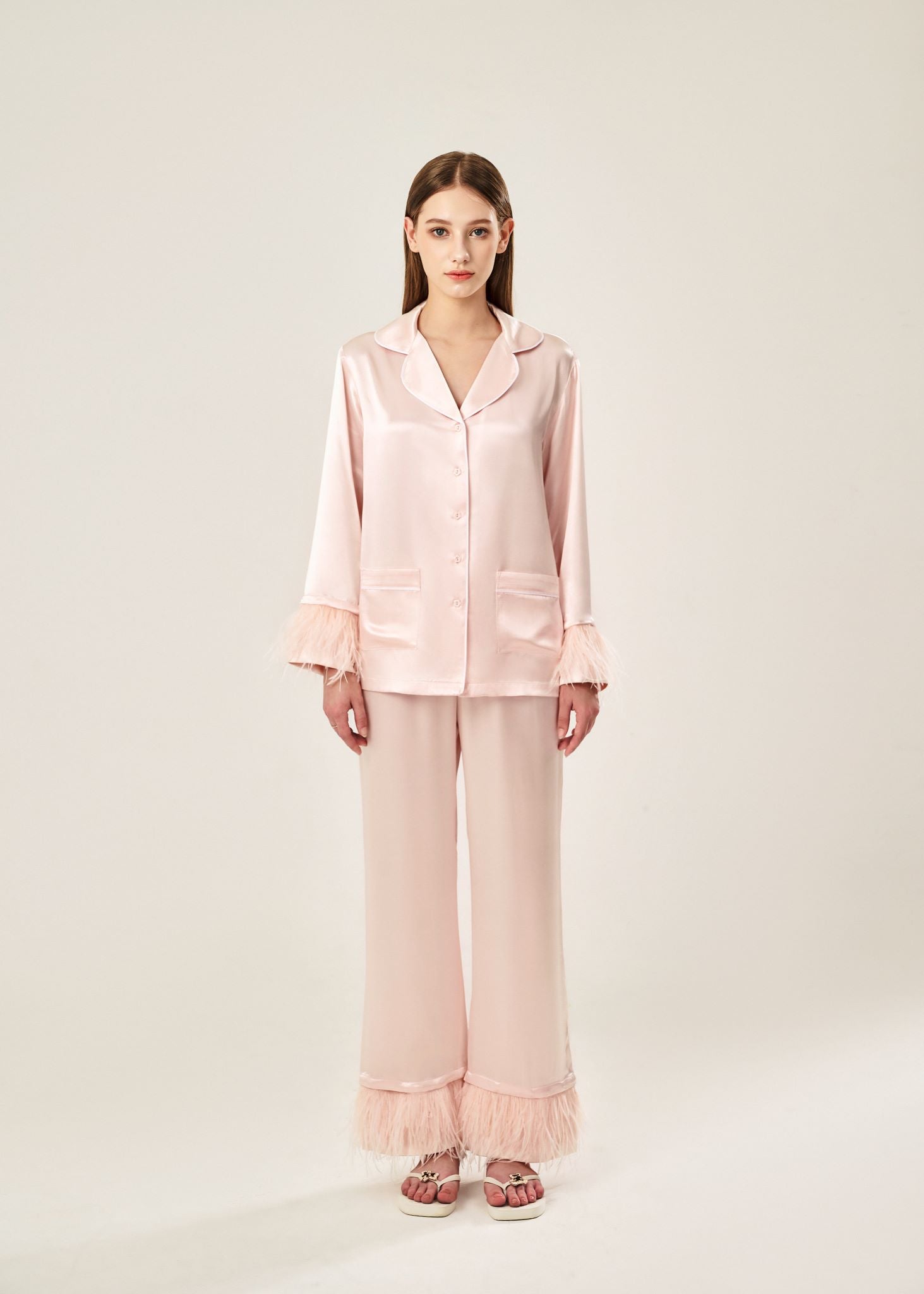 Front view of a blush pink viscose mix rayon pajama set featuring a classic collar, floral buttons, and two front pockets. The detachable ostrich feather trim on the cuffs and pant hems adds a luxurious touch to the relaxed silhouette.