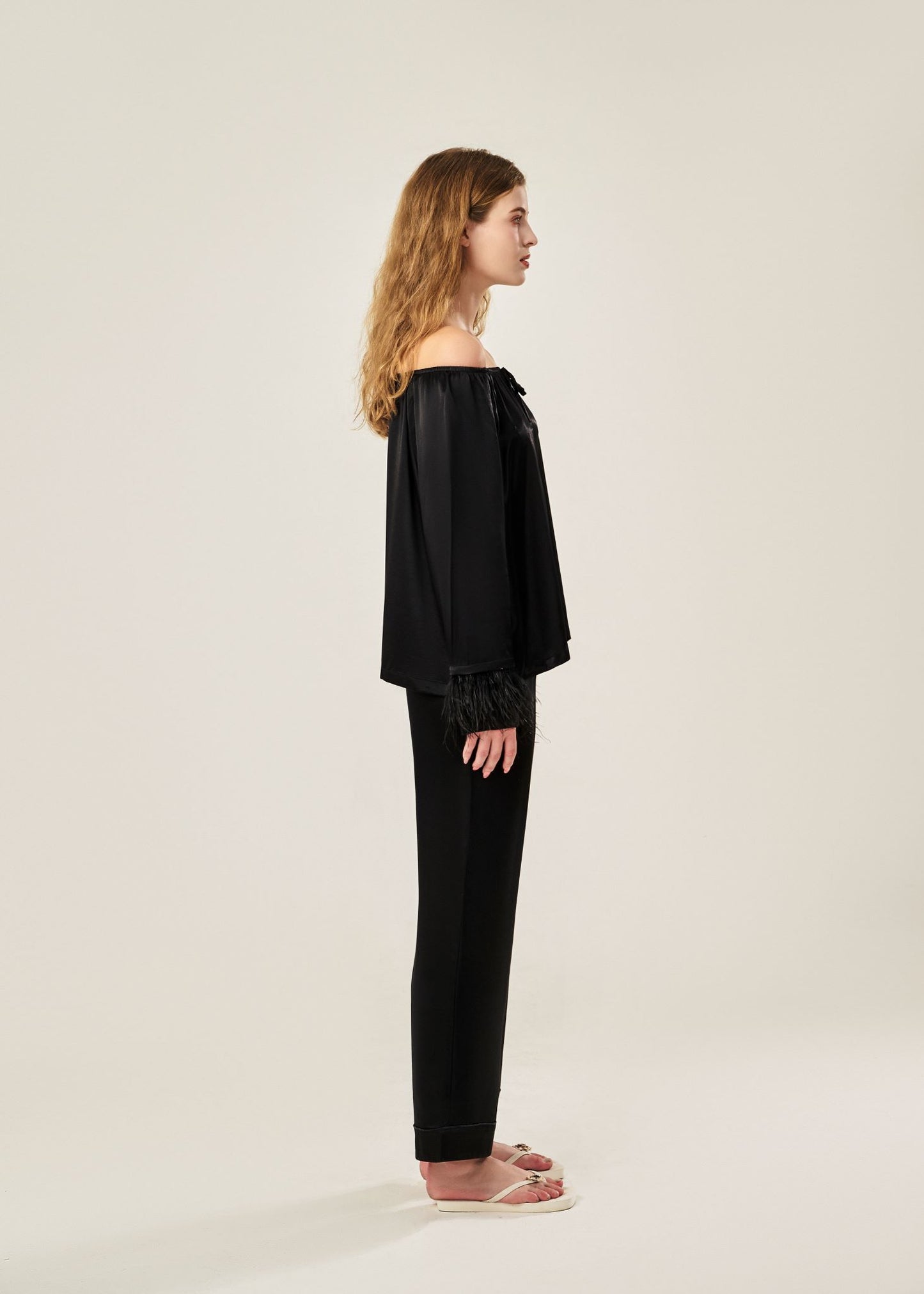 Side view of a black off-shoulder pajama set in soft viscose with a silky feel, featuring a delicate bow detail on the neckline and waistband. Detachable ostrich feather trim on the sleeves adds a chic, elegant finish to the relaxed silhouette.