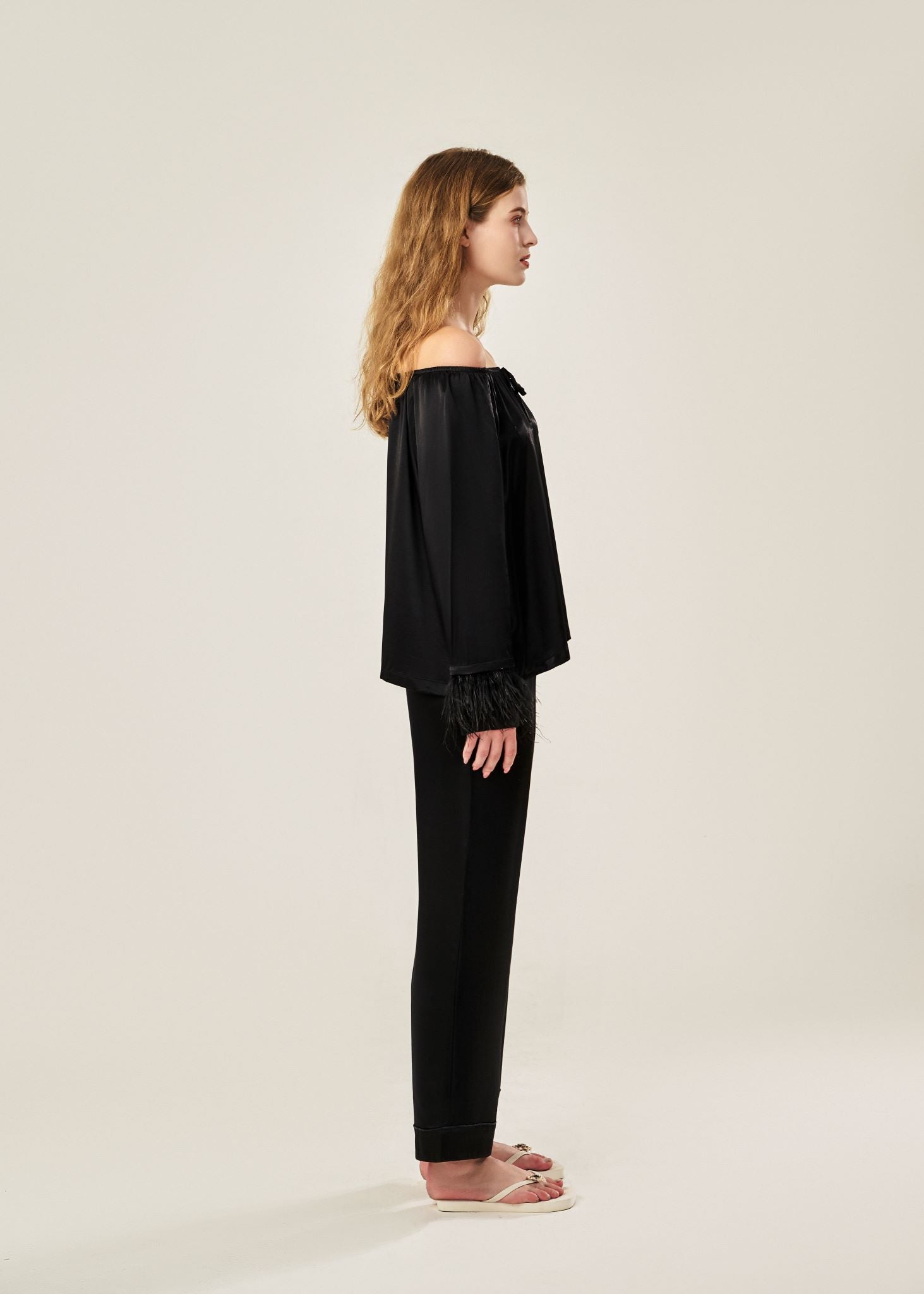 Side view of a black off-shoulder pajama set in soft viscose with a silky feel, featuring a delicate bow detail on the neckline and waistband. Detachable ostrich feather trim on the sleeves adds a chic, elegant finish to the relaxed silhouette.