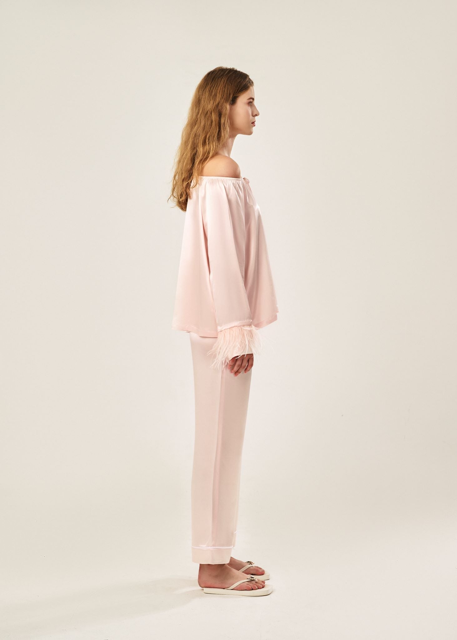 Side view of a blush pink off-shoulder pajama set in soft viscose with a silky feel, featuring a delicate bow detail on the neckline and waistband. Detachable ostrich feather trim on the sleeves adds a chic, elegant finish to the relaxed silhouette.