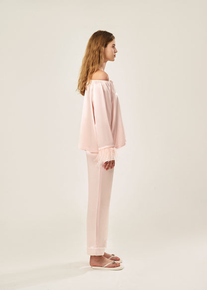 Side view of a blush pink off-shoulder pajama set in soft viscose with a silky feel, featuring a delicate bow detail on the neckline and waistband. Detachable ostrich feather trim on the sleeves adds a chic, elegant finish to the relaxed silhouette.