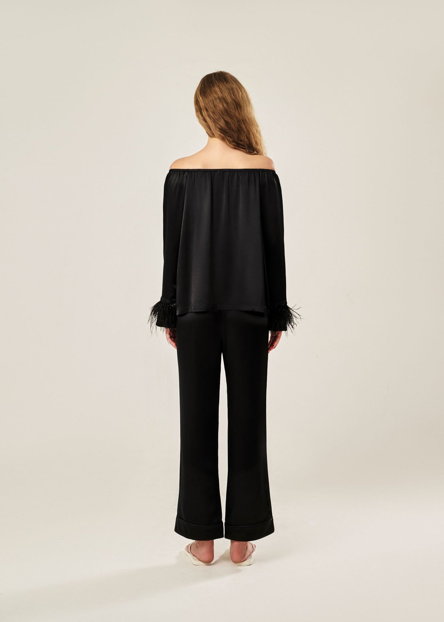 Back view of a black off-shoulder pajama set in soft viscose with a silky feel, featuring a delicate bow detail on the neckline and waistband. Detachable ostrich feather trim on the sleeves adds a chic, elegant finish to the relaxed silhouette.