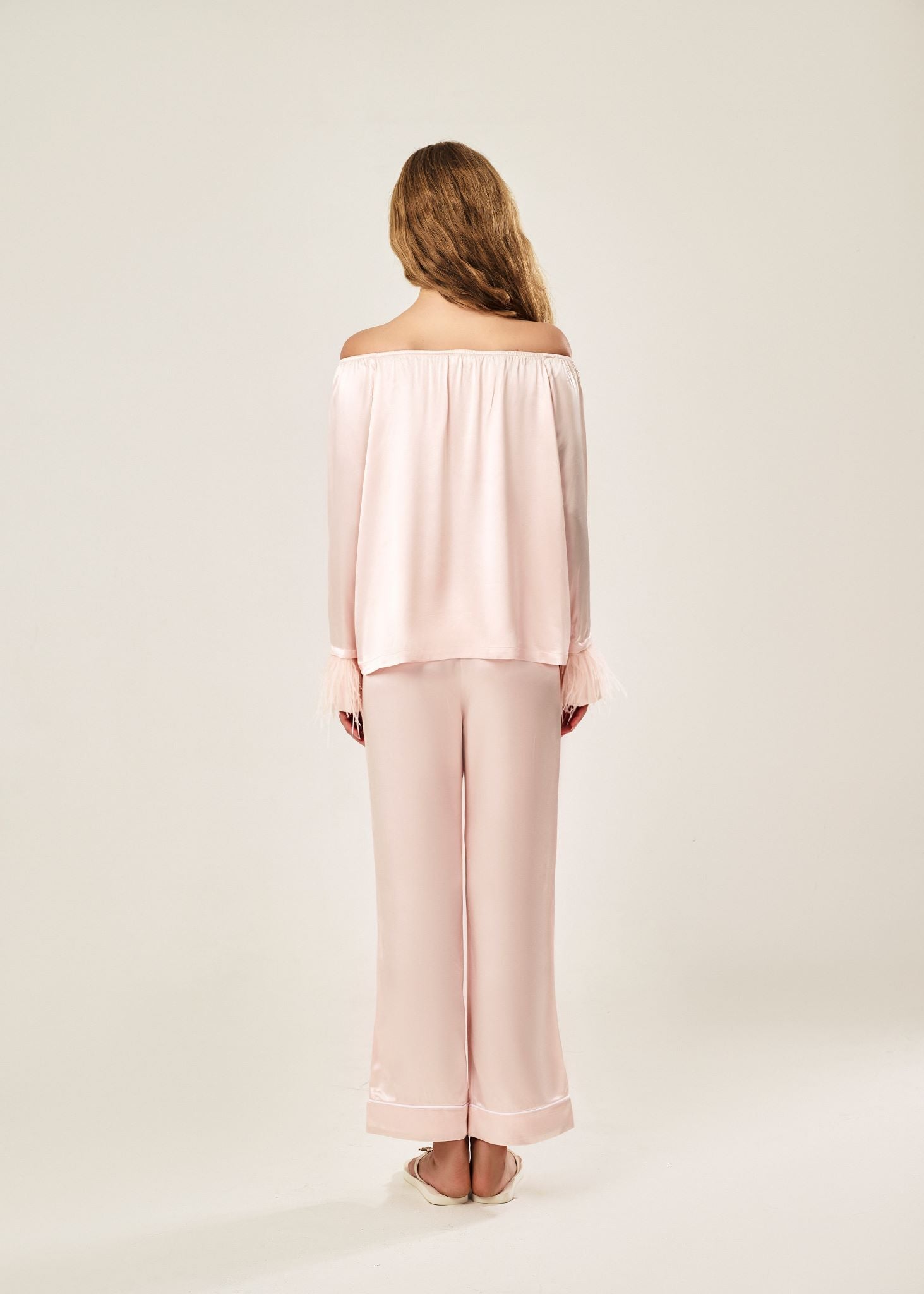 Back view of a blush pink off-shoulder pajama set in soft viscose with a silky feel, featuring a delicate bow detail on the neckline and waistband. Detachable ostrich feather trim on the sleeves adds a chic, elegant finish to the relaxed silhouette.