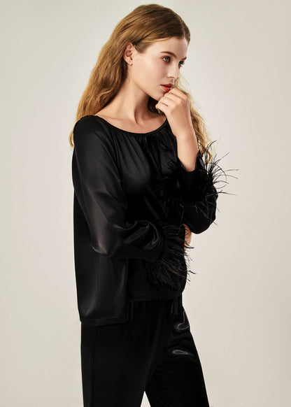 Detail view of a black off-shoulder pajama set in soft viscose with a silky feel, featuring a delicate bow detail on the neckline and waistband. Detachable ostrich feather trim on the sleeves adds a chic, elegant finish to the relaxed silhouette.