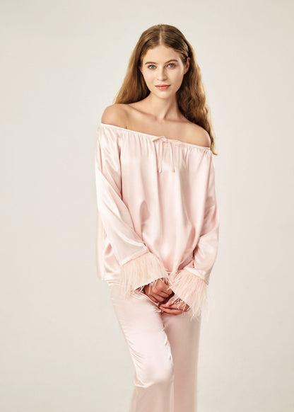 Detail view of a blush pink off-shoulder pajama set in soft viscose with a silky feel, featuring a delicate bow detail on the neckline and waistband. Detachable ostrich feather trim on the sleeves adds a chic, elegant finish to the relaxed silhouette.