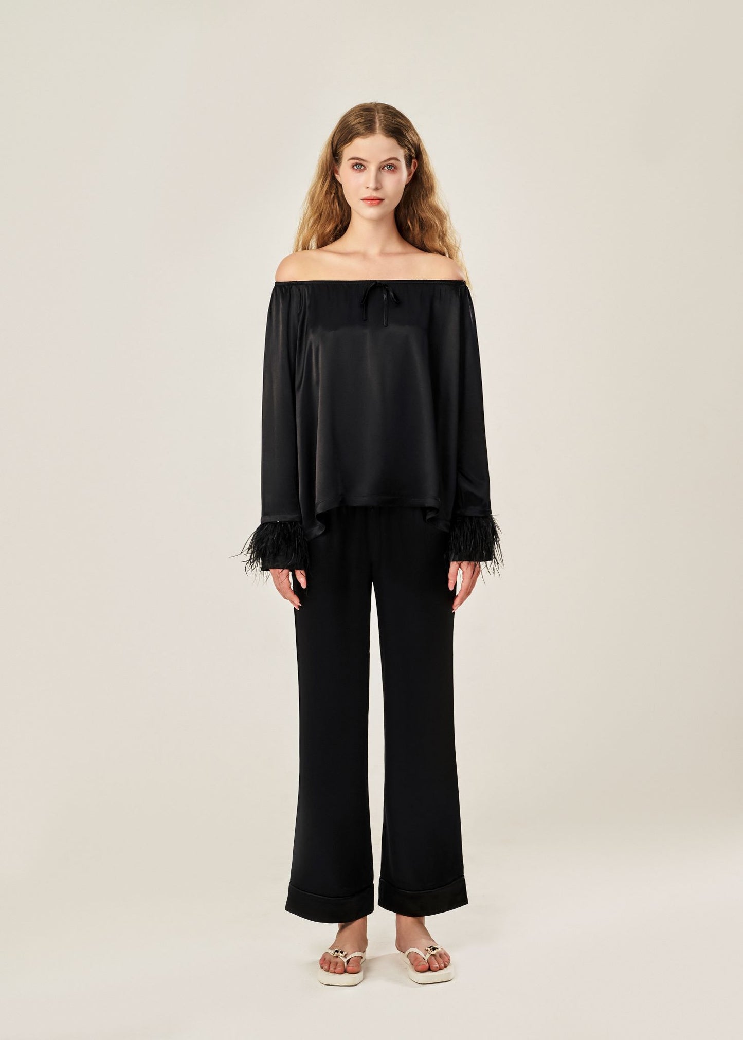 Front view of a black off-shoulder pajama set in soft viscose with a silky feel, featuring a delicate bow detail on the neckline and waistband. Detachable ostrich feather trim on the sleeves adds a chic, elegant finish to the relaxed silhouette.