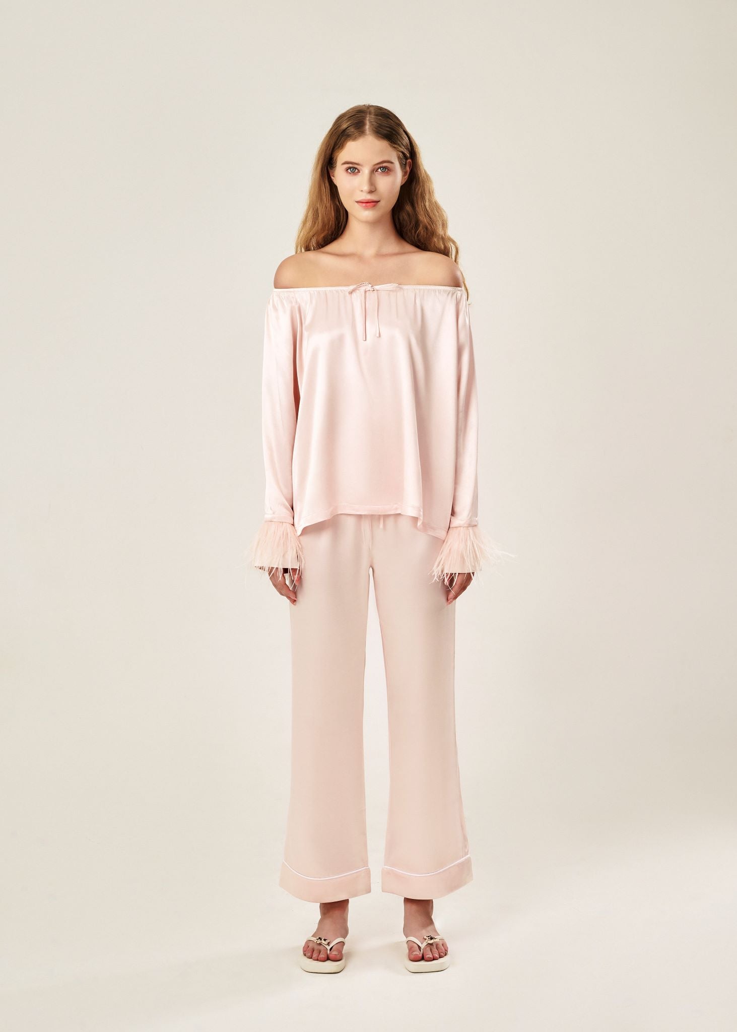 Front view of a blush pink off-shoulder pajama set in soft viscose with a silky feel, featuring a delicate bow detail on the neckline and waistband. Detachable ostrich feather trim on the sleeves adds a chic, elegant finish to the relaxed silhouette.